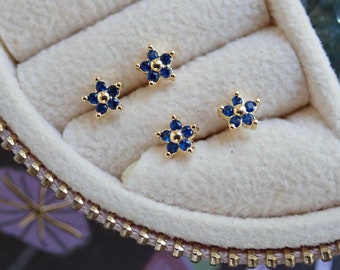 May include: Four gold flower-shaped earrings with blue gemstones in a white velvet jewelry box.