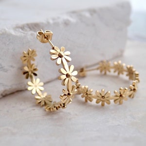 May include: A pair of gold hoop earrings featuring a daisy flower design. The earrings are made of metal and have a polished finish.