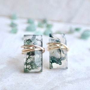 May include: A pair of earrings featuring green moss agate stones wrapped in gold wire. The stones have a translucent white base with green moss-like inclusions.
