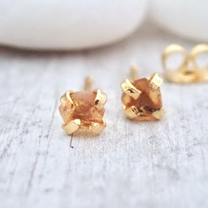 May include: A pair of gold earrings with small, raw, orange-brown gemstones set in a gold claw setting.
