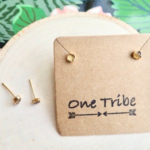 May include: A pair of gold stud earrings with small, raw, light brown gemstones. The earrings are displayed on a brown card with the text "One Tribe" and two arrows.