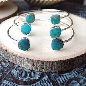 May include: Four silver bangle bracelets with turquoise stones. The bracelets are stacked on top of each other on a wooden surface.