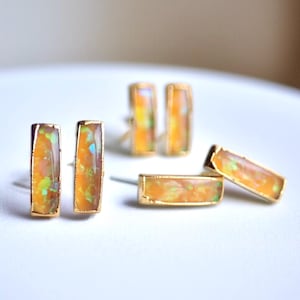 May include: A selection of gold-plated jewellery with rectangular opal stones. The earrings, rings, and studs showcase a warm colour palette with orange, yellow, and green hues. The opals are set in gold frames, creating a luxurious aesthetic.