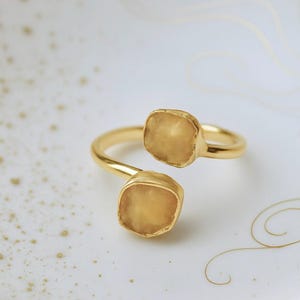 May include: A gold-colored ring with two square, light-yellow gemstones. The ring is set on a white plate with gold-colored decorative lines.