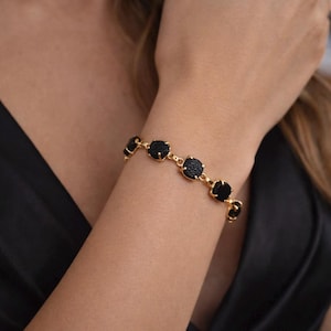 May include: A gold-toned bracelet featuring black, square-shaped stones. The bracelet is worn on a wrist, with the stones evenly spaced and connected by gold links. The background is blurred, focusing attention on the jewelry.