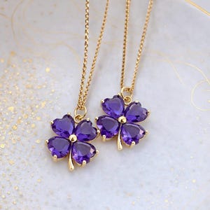 May include: Two gold-colored necklaces, each featuring a four-leaf clover pendant with purple gemstone petals. The pendants rest on a white, gold-rimmed plate with gold flecks.