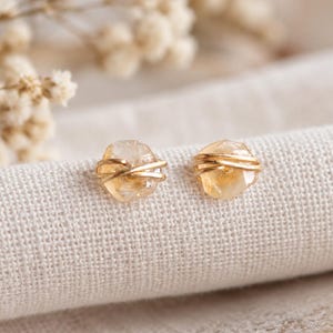 May include: A pair of stud earrings featuring small, faceted, pale yellow citrine gemstones. Each stone is wrapped with thin, polished gold-toned wire that encircles the center, creating a delicate, minimalist design for these gemstone earrings.