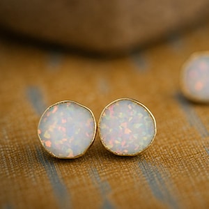 May include: Two pairs of round stud earrings featuring white opalescent stones with iridescent flecks set in thin gold-toned metal bezels. The stones have a smooth, slightly domed surface that reflects light, creating a subtle play of color across the white base.
