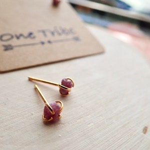 Puede incluir: Un par de pendientes de bot&oacute;n de oro con detalles de piedras preciosas rosas. Los pendientes est&aacute;n colocados sobre una superficie de madera clara. El texto "One Tribe" es visible en una etiqueta de papel marr&oacute;n en el fondo.