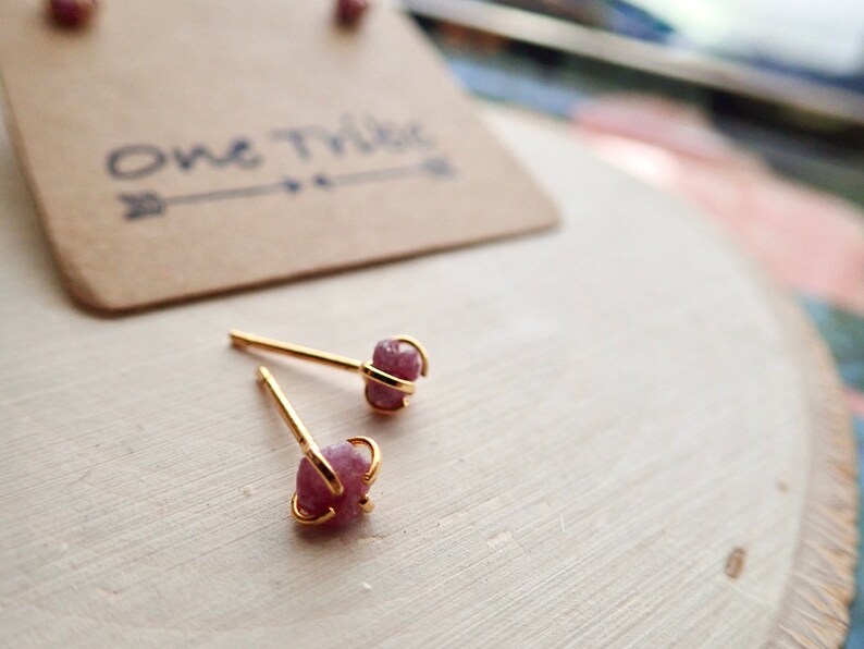 Puede incluir: Un par de pendientes de bot&oacute;n de oro con detalles de piedras preciosas rosas. Los pendientes est&aacute;n colocados sobre una superficie de madera clara. El texto "One Tribe" es visible en una etiqueta de papel marr&oacute;n en el fondo.