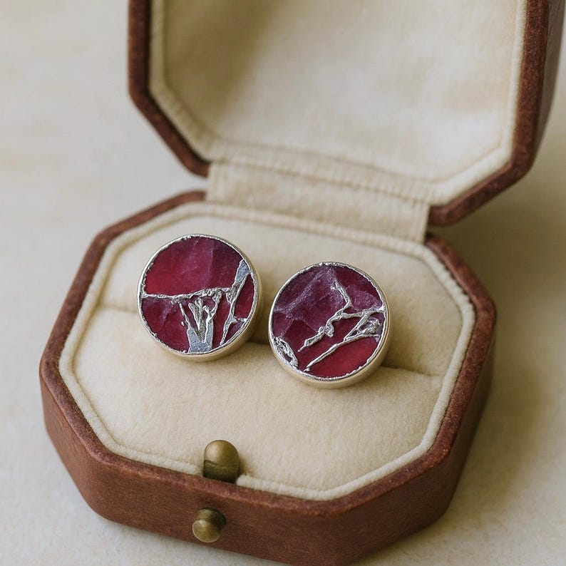 May include: A pair of circular earrings with a silver-coloured metal border. The earrings feature a deep red centre with a silver-coloured, abstract design. They are presented in an open, brown, octagonal jewellery box with a beige interior.