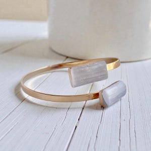 May include: A gold-colored cuff bracelet with two rectangular, light gray crystal accents. The bracelet is open-ended and rests on a white wooden surface. The crystals are a light, translucent gray.