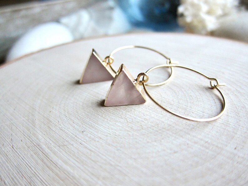 May include: A pair of gold hoop earrings with pink triangle-shaped charms. The charms have a pearlescent finish.