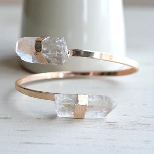 May include: A gold-tone cuff bracelet featuring two clear quartz crystal points. The crystals are secured with gold-colored metal accents. The bracelet has a minimalist design, with a spiral shape.