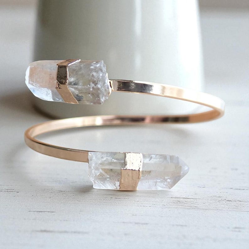 May include: A gold-tone cuff bracelet featuring two clear quartz crystal points. The crystals are secured with gold-colored metal accents. The bracelet has a minimalist design, with a spiral shape.