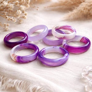 May include: A collection of gemstone rings in shades of purple, lavender, and white. The rings are smooth and polished, with varying patterns and color gradients. The rings are displayed on a white fabric surface.