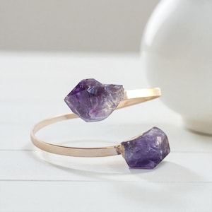 May include: A gold-toned open cuff bracelet featuring two raw, deep purple amethyst crystals at each end. The band is smooth and metallic, curving into a circular shape with the two jagged, natural-cut stones facing each other at the opening.