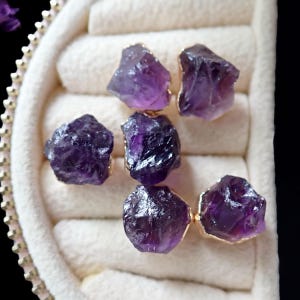 May include: Six raw amethyst stones with gold-colored edges are arranged in a jewelry box. The amethyst crystals display a deep purple hue, with a rough, natural texture. The gold edges add a touch of elegance to the gemstones.