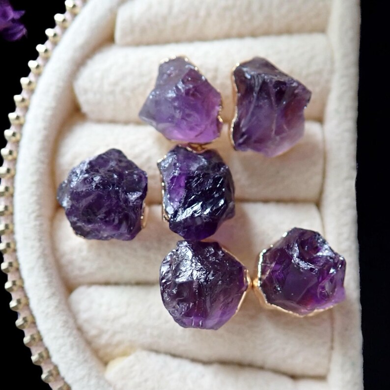 May include: Six raw amethyst stones with gold-colored edges are arranged in a jewelry box. The amethyst crystals display a deep purple hue, with a rough, natural texture. The gold edges add a touch of elegance to the gemstones.