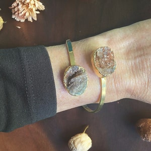 May include: A gold bangle bracelet with two attached, natural, brown and white agate stones. The bracelet is on a person's wrist.