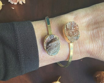 May include: A gold bangle bracelet with two attached, natural, brown and white agate stones. The bracelet is on a person's wrist.