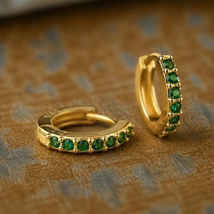 May include: A pair of gold hoop earrings featuring a row of small, round, vibrant green gemstones set along the front edge of each hoop. The earrings have a polished metallic finish and a hinged closure mechanism for secure wear.