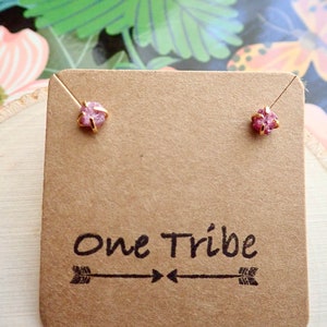 Puede incluir: Un par de pendientes con peque&ntilde;as piedras preciosas rosas engastadas en oro. Los pendientes se muestran en una tarjeta marr&oacute;n con el texto "One Tribe" y un dise&ntilde;o de flecha.