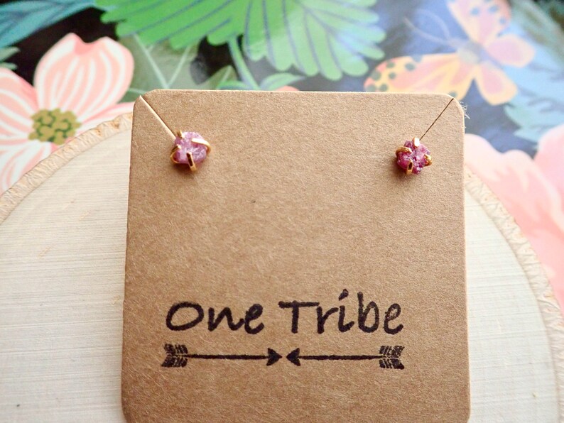 Puede incluir: Un par de pendientes con peque&ntilde;as piedras preciosas rosas engastadas en oro. Los pendientes se muestran en una tarjeta marr&oacute;n con el texto "One Tribe" y un dise&ntilde;o de flecha.