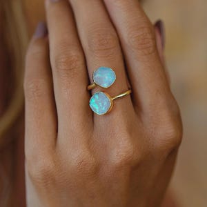 May include: A gold-colored ring featuring two oval, light blue opal stones. The ring is worn on a hand, showcasing the gemstones' iridescent quality. The ring's design is simple, with the stones set in a bezel setting.
