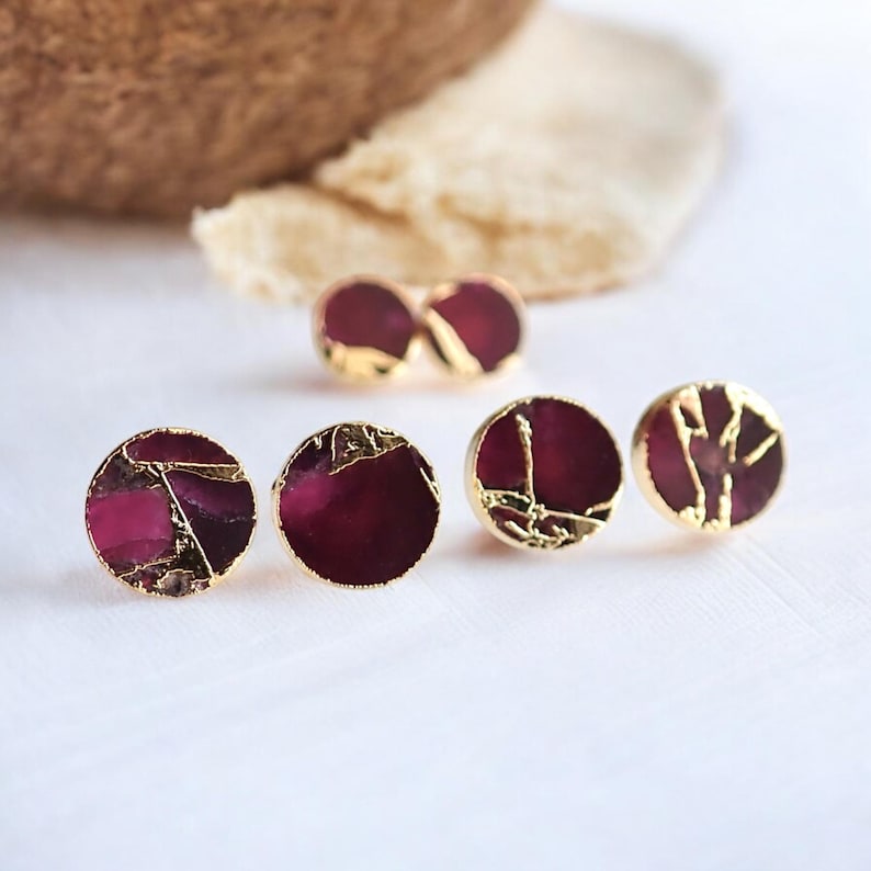 May include: Gold-edged stud earrings with a rich burgundy centre. The earrings have a distinctive, textured look with gold detailing. The image displays several pairs of earrings, emphasising their design and colour.