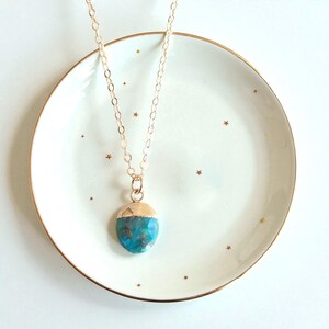 Peut inclure: Un collier &agrave; cha&icirc;ne dor&eacute;e avec un pendentif turquoise. Le pendentif est de forme arrondie avec un capuchon dor&eacute;. Le collier repose sur une assiette blanche avec un bord dor&eacute; et de petites &eacute;toiles dor&eacute;es.