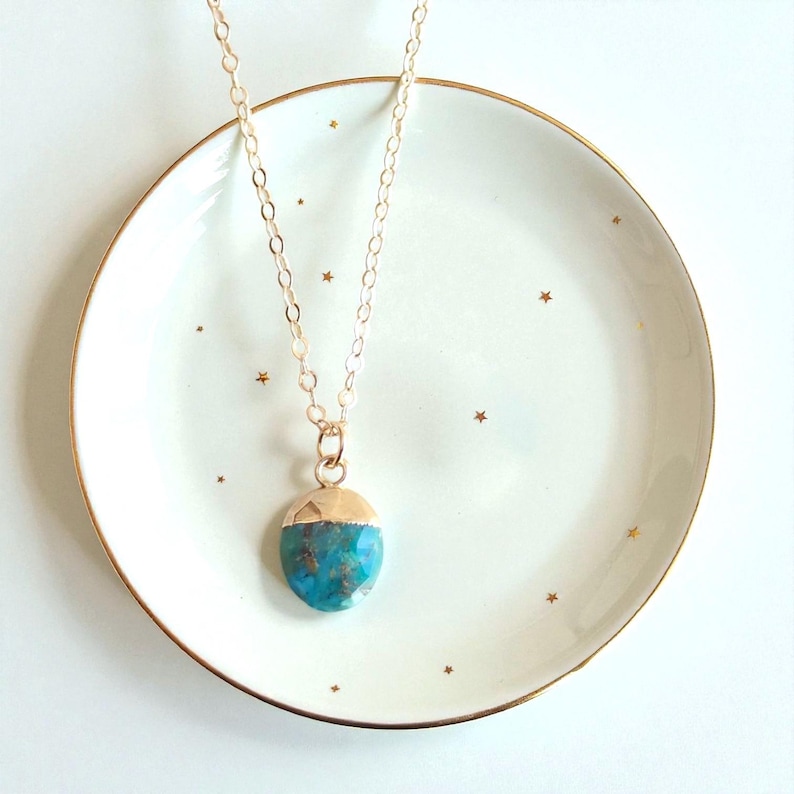 Peut inclure: Un collier &agrave; cha&icirc;ne dor&eacute;e avec un pendentif turquoise. Le pendentif est de forme arrondie avec un capuchon dor&eacute;. Le collier repose sur une assiette blanche avec un bord dor&eacute; et de petites &eacute;toiles dor&eacute;es.