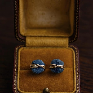 Può includere: Un paio di orecchini in pietra blu avvolti in filo d'oro, esposti in un portagioie bordeaux vintage aperto con interno dorato. Gli orecchini sono il punto focale, mostrando un design minimalista.