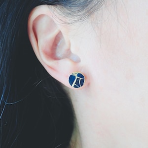 May include: A close-up of a dark blue and gold stud earring. The round earring has a gold border and a dark blue centre with a gold, cracked pattern. The earring is worn on a person's ear.