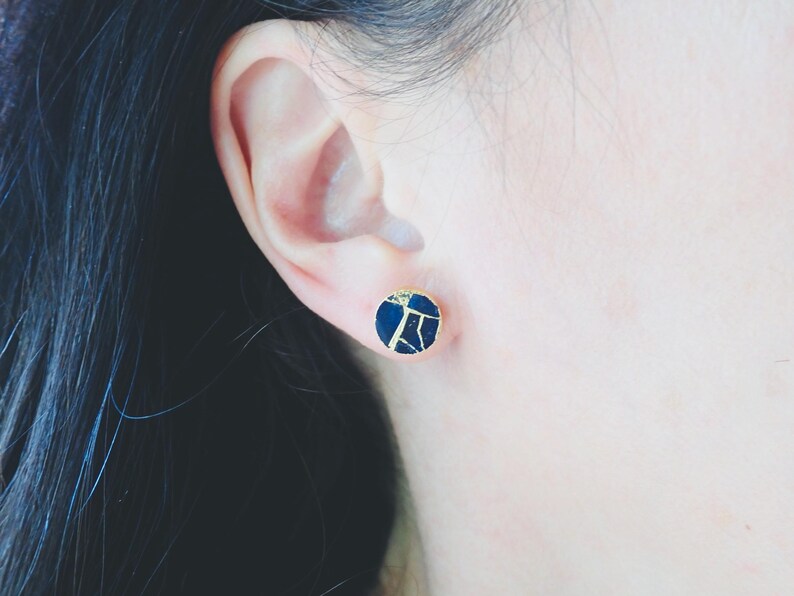 May include: A close-up of a dark blue and gold stud earring. The round earring has a gold border and a dark blue centre with a gold, cracked pattern. The earring is worn on a person's ear.