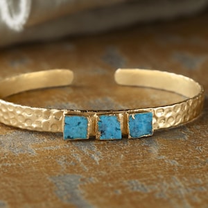 May include: A gold-toned cuff bracelet featuring a hammered metal texture and three small, square-shaped, bright blue turquoise stones set in a row along the center of the band. The bracelet has an open back design for easy adjustment.