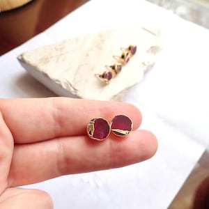May include: Round stud earrings with a gold trim and a deep red gemstone centre. Additional earrings with the same design are visible in the background, resting on a white stone.