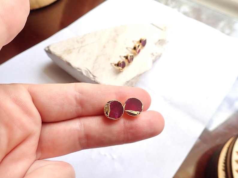 May include: Round stud earrings with a gold trim and a deep red gemstone centre. Additional earrings with the same design are visible in the background, resting on a white stone.