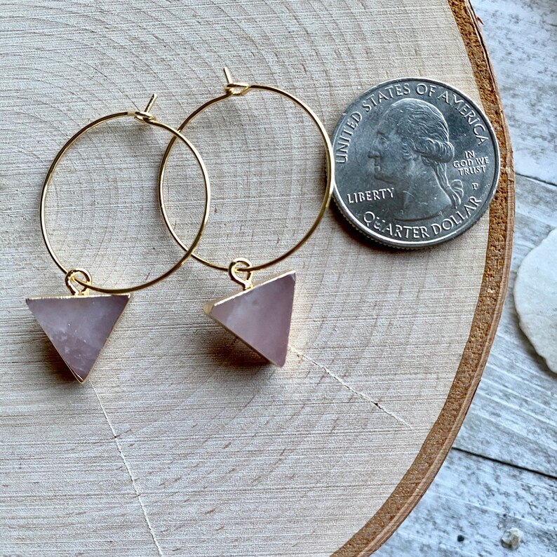 May include: A pair of gold hoop earrings with pink triangle-shaped charms. The earrings are shown next to a US quarter for size reference.