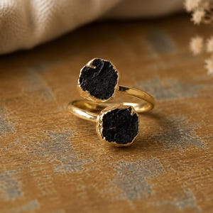 May include: A gold-toned bypass ring featuring two raw, black, textured stones set at each end of the open band. The stones have a rough, irregular shape and a matte finish, contrasting with the smooth, polished surface of the metallic ring band.
