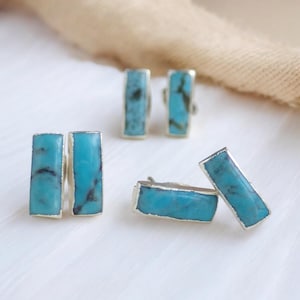 May include: A collection of turquoise and silver earrings. Each earring features a rectangular turquoise stone, with dark blue and black veining, set in a silver frame. The earrings are displayed on a white surface.