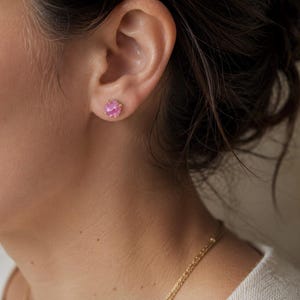 May include: A single stud earring featuring a round, faceted pink gemstone set in a gold-toned metal prong setting. The earring is positioned in the earlobe, showcasing the vibrant pink colour of the stone and the metallic finish of the prongs.
