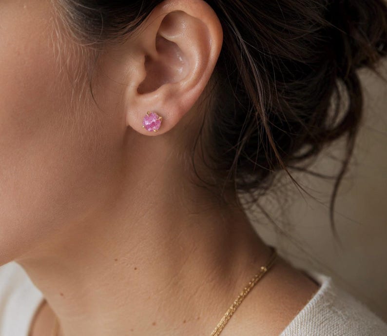 May include: A single stud earring featuring a round, faceted pink gemstone set in a gold-toned metal prong setting. The earring is positioned in the earlobe, showcasing the vibrant pink colour of the stone and the metallic finish of the prongs.