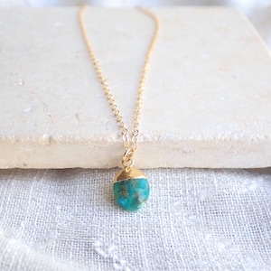 Peut inclure: Un collier &agrave; cha&icirc;ne dor&eacute;e avec un pendentif turquoise. Le pendentif a une forme brute et irr&eacute;guli&egrave;re avec un capuchon dor&eacute;. Le collier est pr&eacute;sent&eacute; sur une surface blanche.