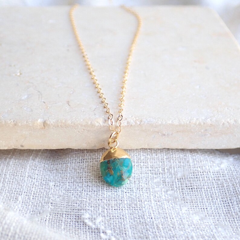 Peut inclure: Un collier &agrave; cha&icirc;ne dor&eacute;e avec un pendentif turquoise. Le pendentif a une forme brute et irr&eacute;guli&egrave;re avec un capuchon dor&eacute;. Le collier est pr&eacute;sent&eacute; sur une surface blanche.