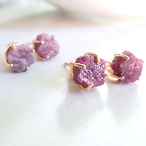 May include: Two pairs of stud earrings featuring raw, deep pink gemstone crystals. Each rough-cut stone is secured in a gold-toned metal prong setting. The earrings are arranged in a cluster, showcasing the irregular, textured surfaces of the stones.