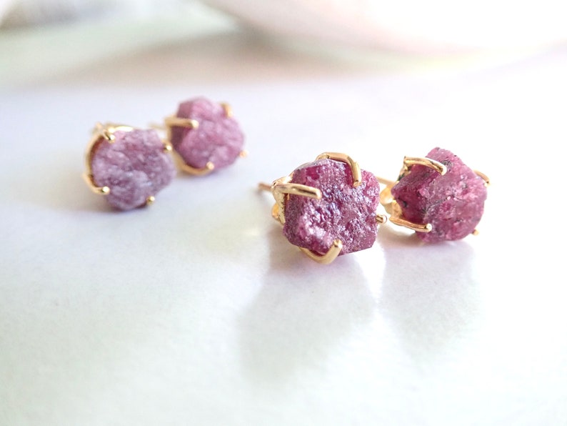 May include: Two pairs of stud earrings featuring raw, deep pink gemstone crystals. Each rough-cut stone is secured in a gold-toned metal prong setting. The earrings are arranged in a cluster, showcasing the irregular, textured surfaces of the stones.