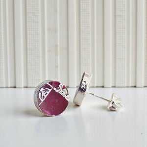 May include: A pair of silver stud earrings. Each earring has a circular shape with a deep pink centre and a textured silver border. The earrings are displayed on a white surface, highlighting their design and craftsmanship.