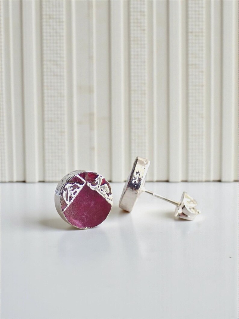 May include: A pair of silver stud earrings. Each earring has a circular shape with a deep pink centre and a textured silver border. The earrings are displayed on a white surface, highlighting their design and craftsmanship.