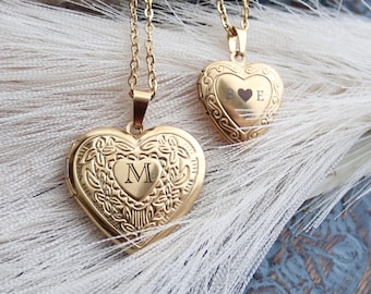 May include: Two gold heart-shaped locket necklaces. The locket on the left has the initial "M" engraved on it. The locket on the right has the initials "B" and "E" engraved on it, with a small heart between the letters.